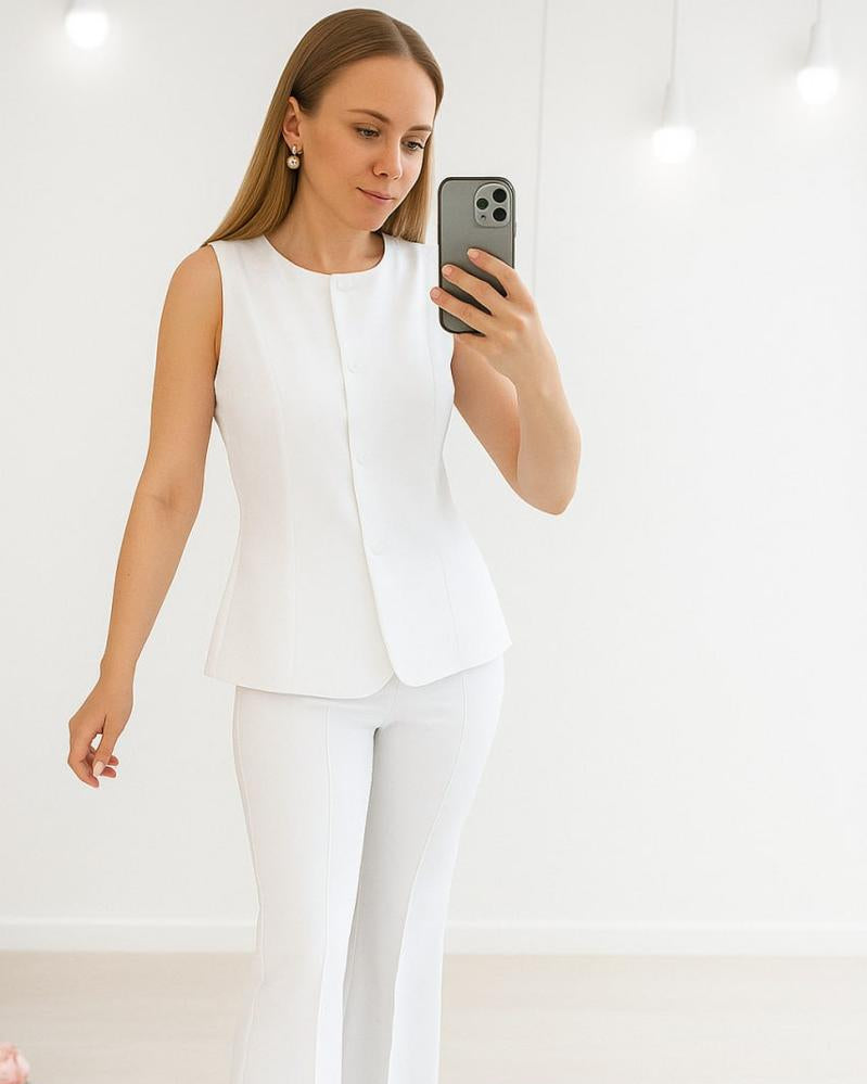 Woman taking a mirror selfie wearing a white sleeveless blazer and pants.
