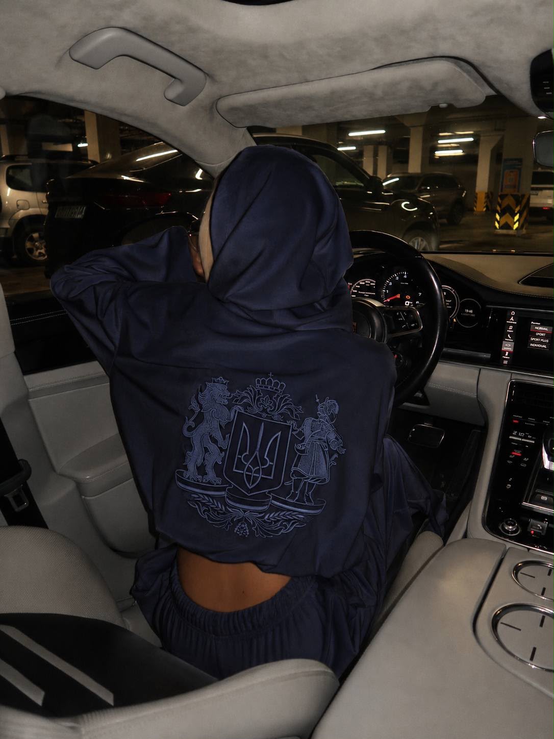 Woman wearing dark blue velour hoodie with Ukrainian coat of arms embroidery, hood up, back view in car