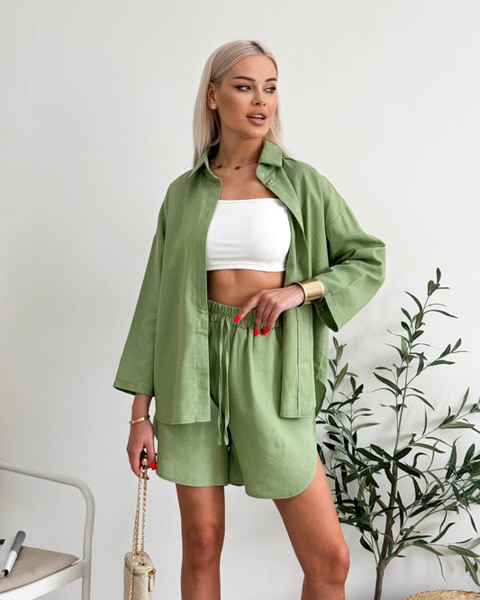 Woman wearing a green outfit with a white top, standing indoors with a plant in the background.