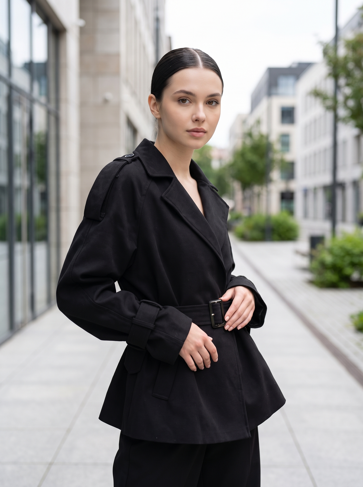 Woman wearing black suede-finish belted trench coat with epaulettes, front three-quarter view on city street